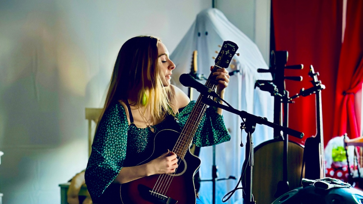 Darcy Rose Byrnes, a young caucasion woman with long blonde hair, plays a guitar and sings into a microphone. A sheet ghost stands a few feet behind her.